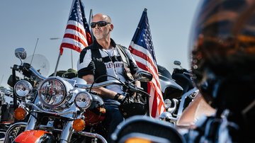 Rider sits on parked bike with United State flags in the background